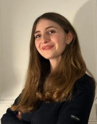 Portrait d’une jeune femme souriante aux longs cheveux bruns, portant un haut noir, photographiée contre un mur clair.