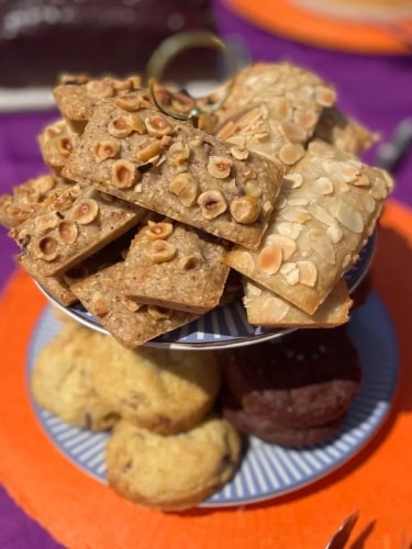 Un présentoir à deux étages garni de financiers aux noisettes et aux amandes, accompagnés de biscuits ronds nature et au chocolat sur une nappe orange et violette.