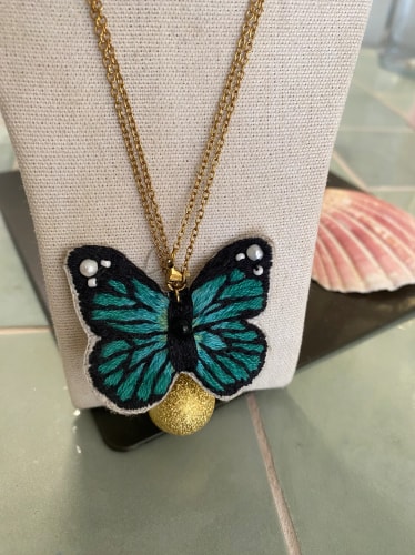 Collier avec un pendentif papillon brodé en bleu turquoise et noir, orné de petites perles blanches, sur une chaîne dorée, présenté sur un support en tissu beige avec un coquillage en arrière-plan.