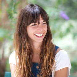 Jeune femme souriante aux longs cheveux bruns, portant une blouse blanche et une salopette bleue, dans un jardin verdoyant.