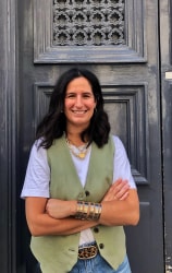 Femme souriante aux cheveux noirs, vêtue d’un gilet vert et d’un haut lavande, bras croisés, devant une porte noire ornée.