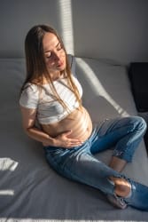 Une femme enceinte noire avec des dreadlocks, assise en tailleur contre un mur blanc, portant un haut noir, un gilet en jean sans manches et des bijoux, le regard tourné sur le côté en souriant.