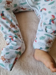 Jambes d'un bébé portant un pyjama blanc à motifs de singes et de toucans, l'un des pieds est couvert et l'autre est nu sur une couverture beige douce.