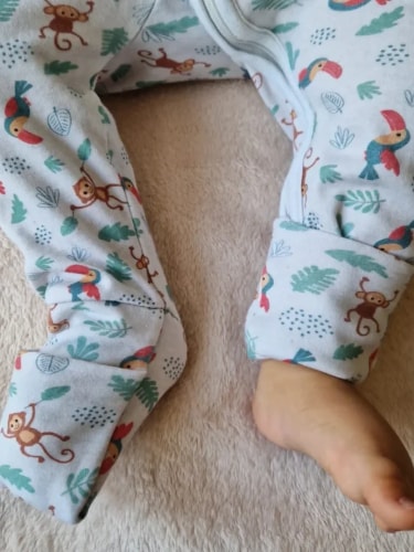 Jambes d'un bébé portant un pyjama blanc à motifs de singes et de toucans, l'un des pieds est couvert et l'autre est nu sur une couverture beige douce.