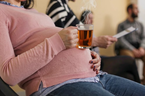 Femme enceinte portant un pull rose, tenant une tasse de thé fumante sur son ventre, assise dans une salle d'attente avec d'autres personnes en arrière-plan.