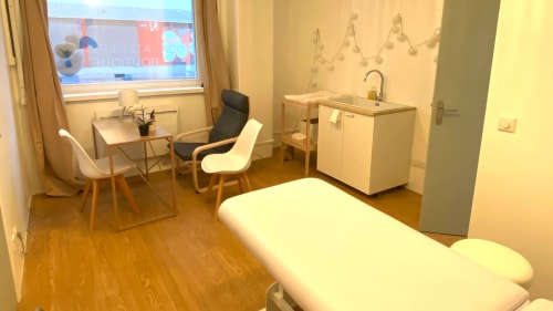 Une salle de massage minimaliste et chaleureuse avec une table de soin, un coussin orange, une serviette à franges, et un buffet blanc décoré d'une plante sous une fresque murale abstraite.