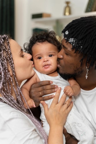 Femme enceinte blonde portant un débardeur noir et une veste à motifs, tenant délicatement un petit cœur en tissu rayé contre son ventre dénudé, avec un sourire bienveillant.