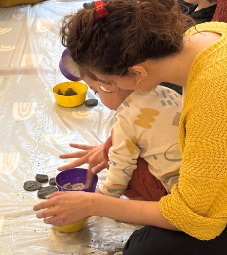 Une femme en pull jaune joue avec un jeune enfant, manipulant de la terre ou de la pâte dans des bols colorés posés sur une nappe en plastique sur le sol.