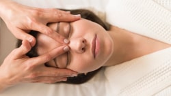 Une femme allongée dans un peignoir blanc reçoit un massage du visage relaxant par une esthéticienne qui masse délicatement la zone entre ses sourcils et ses tempes.