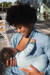 Femme enceinte blonde portant un débardeur noir et une veste à motifs, tenant délicatement un petit cœur en tissu rayé contre son ventre dénudé, avec un sourire bienveillant.