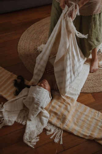 Une personne étendue sur le sol est recouverte de tissus beiges et blancs par quelqu'un, dans une ambiance douce sur un tapis en paille.