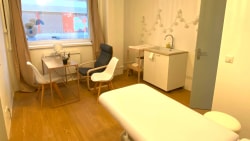 Cabinet de consultation propre avec une table d'examen blanche, un bureau avec des chaises, un lavabo, des rangements et deux suspensions lumineuses au plafond. Fenêtre avec rideaux beiges.