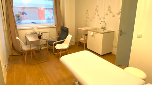 Cabinet de consultation propre avec une table d'examen blanche, un bureau avec des chaises, un lavabo, des rangements et deux suspensions lumineuses au plafond. Fenêtre avec rideaux beiges.