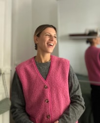 Femme souriante portant un gilet rose vif sur un pull gris, debout dans une pièce lumineuse, avec son reflet visible dans un miroir.