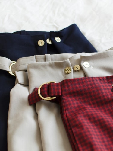 Trois pantalons à taille haute superposés, aux couleurs bleu marine, beige et rouge à carreaux, chacun doté d'une ceinture à boucle métallique dorée et de boutons.