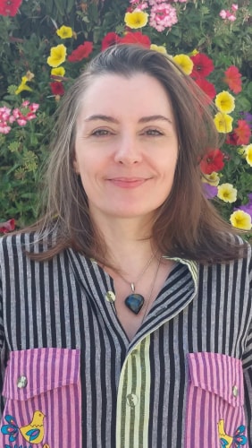 Portrait d’une femme souriante aux cheveux longs, devant un mur fleuri, portant une chemise rayée à poches colorées et un pendentif.