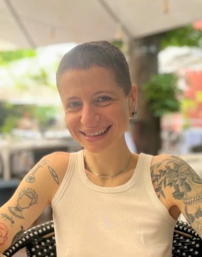 Portrait souriant d'une personne aux cheveux courts, portant un débardeur blanc et des tatouages sur les bras, assise à une terrasse en arrière-plan flou.