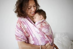 Femme enceinte blonde portant un débardeur noir et une veste à motifs, tenant délicatement un petit cœur en tissu rayé contre son ventre dénudé, avec un sourire bienveillant.