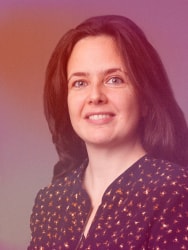 Portrait d’une femme souriante aux cheveux roux foncé, vêtue d’un chemisier sombre à motifs dorés, sur un fond dégradé rose et violet.