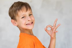 Jeune garçon souriant vêtu d'un t-shirt orange, faisant un geste de la main indiquant que tout va bien, sur fond gris.