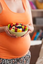 Une femme enceinte portant un débardeur orange tient un bol rempli de salade de fruits frais devant son ventre.