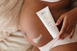 Une femme enceinte noire pose avec un tube de lait corporel Daylily posé sur son ventre, où une petite noisette de crème est appliquée.