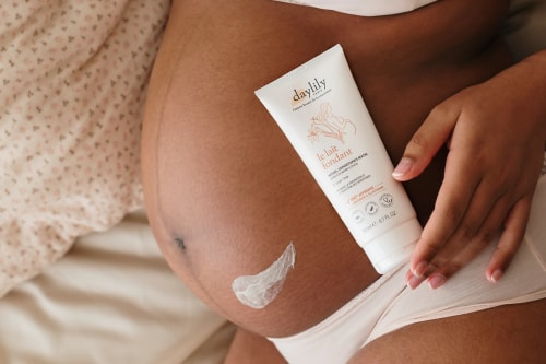 Une femme enceinte noire pose avec un tube de lait corporel Daylily posé sur son ventre, où une petite noisette de crème est appliquée.