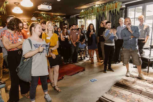 Groupe de personnes souriantes assistant à un événement en intérieur, certaines tenant des dossiers et des téléphones, avec une ambiance conviviale et un décor végétal suspendu.