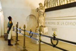 Un soldat en uniforme d'époque regarde une statue en pierre d'un soldat avec un fusil dans un monument commémoratif. L'inscription au mur indique « SOLDAT INCONNU DE LA GRANDE ».
