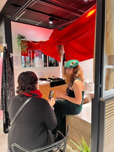 Une femme assise de dos tenant une carte, en face d'une autre femme portant un chapeau vert avec un motif, assise sur le bord d'une baignoire dans une pièce décorée avec un tissu rouge suspendu et des plantes.