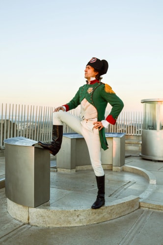 Homme en costume historique de style Napoléonien vert et blanc avec un chapeau bicorne noir, posant avec une jambe levée sur un socle en pierre dans un espace extérieur moderne avec des barrières métalliques