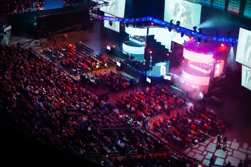 Un homme portant un masque bleu et une cape posant avec un ordinateur portable tenu dans la main, tandis que trois femmes l'applaudissent dans un environnement intérieur avec des plantes vertes.