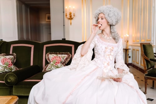 Femme habillée en costume d'époque baroque avec une perruque blanche bouclée, assise sur un canapé vert avec coussins fleuris, en train de manger un macaron dans un intérieur élégant avec des murs blancs et dorés.