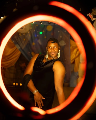 Personne posant dans une robe noire et portant de grands boucles d'oreilles brillantes, encadrée par un cercle lumineux rouge orangé.