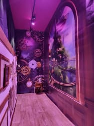 Couloir décoré avec des illustrations de rouages d'horlogerie et d'horloges, ambiance lumineuse violette, avec un petit meuble et un tableau noir à l'extrémité.