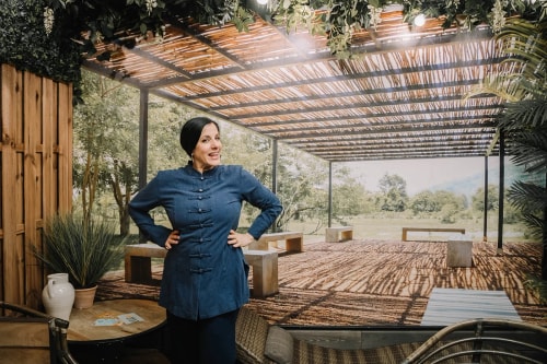 Femme souriante vêtue d'une tunique bleue foncée, debout avec les mains sur les hanches dans un espace intérieur décoré avec des plantes, une table en bois avec des cartes, et un grand mur avec une image représentant une terrasse ombragée en plein air entourée de nature.