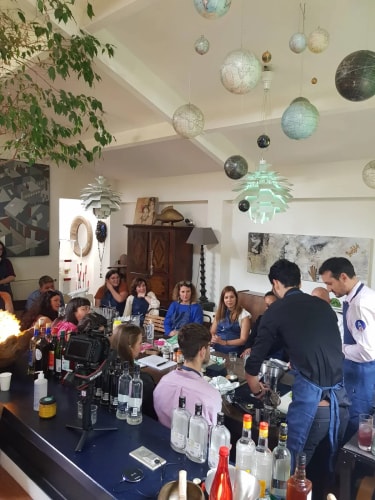 Groupe de personnes assises autour d'une table dans une pièce, deux hommes debout préparent des boissons, plusieurs bouteilles sur la table, décor de globes suspendus au plafond.