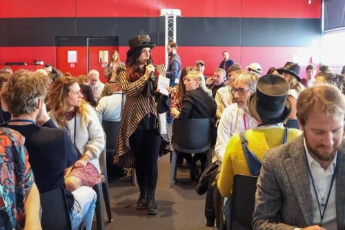 Frances Vieras-Blanc debout au centre portant un chapeau et tenant un microphone, s'adressant à un groupe de personnes assises dans une salle avec un mur rouge et noir en arrière-plan.