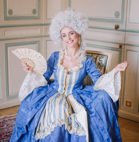 Femme souriante en costume d'époque bleu et blanc avec une perruque grise bouclée, tenant un éventail, assise sur une chaise dans une pièce ornée de boiseries.