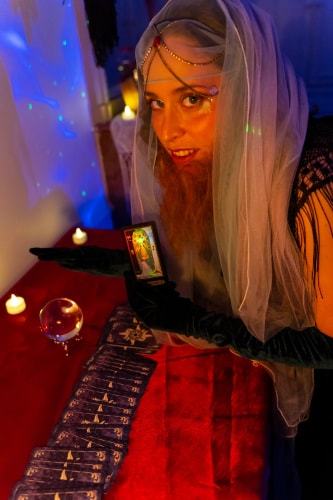 Une voyante avec un voile transparent et des gants noirs, montrant une carte de tarot dans une pièce faiblement éclairée avec des bougies allumées et une boule de cristal sur une table recouverte de tissu rouge et une rangée de cartes de tarot étalées.
