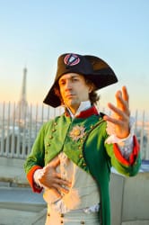 Un homme en costume d'époque napoléonienne avec un bicorne noir orné d'un cocarde tricolore, une veste verte à boutons dorés, posant avec une main sur le ventre et l'autre tendue vers l'avant, avec la tour Eiffel en arrière-plan au coucher du soleil.