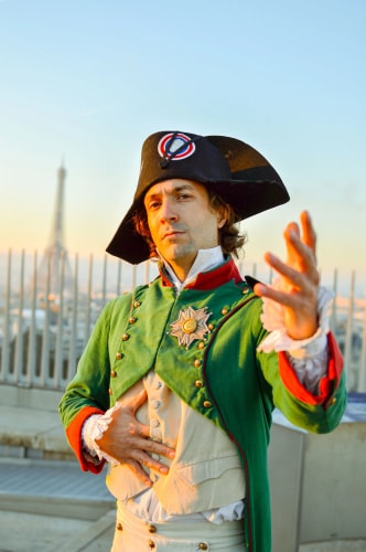 Un homme en costume d'époque napoléonienne avec un bicorne noir orné d'un cocarde tricolore, une veste verte à boutons dorés, posant avec une main sur le ventre et l'autre tendue vers l'avant, avec la tour Eiffel en arrière-plan au coucher du soleil.