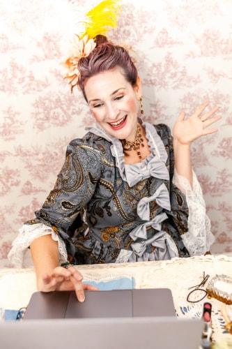 Une femme souriante vêtue d'une robe ancienne ornée de nœuds gris et de dentelle, coiffée avec une plume jaune et une fleur orange, interagit avec un ordinateur portable devant elle, sur une table recouverte d'une nappe en dentelle.