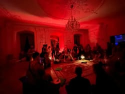 Groupe de personnes assises en cercle dans une grande pièce décorée, éclairée par une lumière rouge avec un grand lustre au plafond.