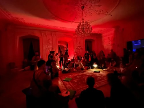 Groupe de personnes assises en cercle dans une grande pièce décorée, éclairée par une lumière rouge avec un grand lustre au plafond.