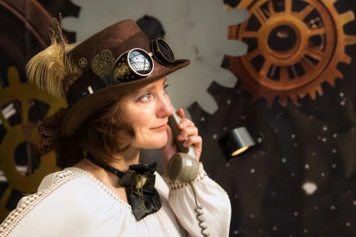 Une femme habillée en style steampunk avec un chapeau orné de plumes et de lunettes, parlant au téléphone ancien, devant un fond avec des engrenages géants.