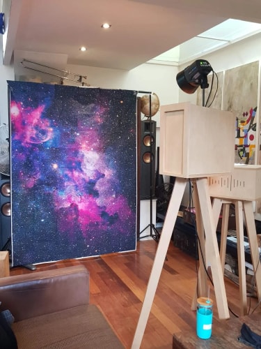 Une pièce avec un mur décoré d'une grande image d'une nébuleuse colorée en rose et bleu, un projecteur noir monté sur un trépied en bois clair, des enceintes, un globe terrestre, et un canapé en cuir marron au premier plan.