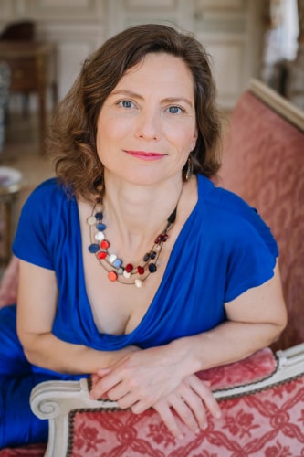 Portrait photo d'Elena Odessa Ray, qui a co-construit Eat the Cake. Elle est assise sur un fauteuil, porte un haut bleu et sourit.