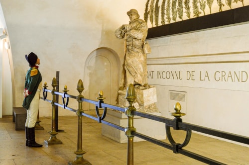 Un homme en costume militaire historique regarde une statue en pierre représentant un soldat avec un uniforme et un casque, située dans un intérieur avec une barrière en métal doré et noir, avec une inscription murale en arrière-plan mentionnant le soldat inconnu de la Grande Guerre.
