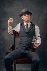 Homme assis sur une chaise, portant un costume gris avec une cravate et un chapeau, tenant une bouteille et un verre de whisky, avec un pistolet attaché au dossier de la chaise, sur un fond sombre.